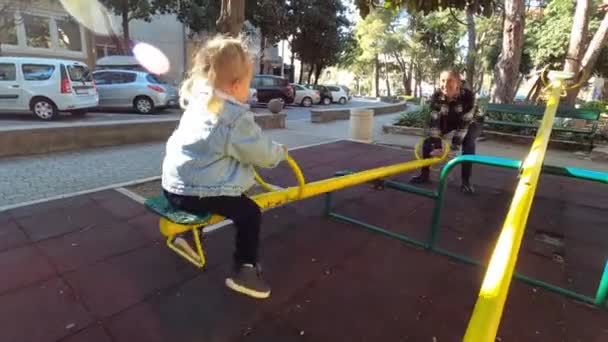 Мама трусить маленьку дівчинку на гойдалці-балансувальнику на ігровому майданчику у дворі будинку. Кадри високої якості 4k - Кадри, відео