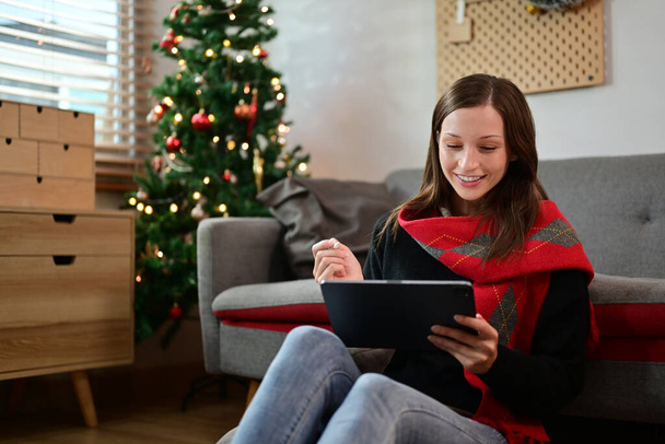 Valkoihoinen nainen freelancer käyttäen digitaalinen tabletti istuessaan olohuoneessa kaunis joulukuusi. - Valokuva, kuva