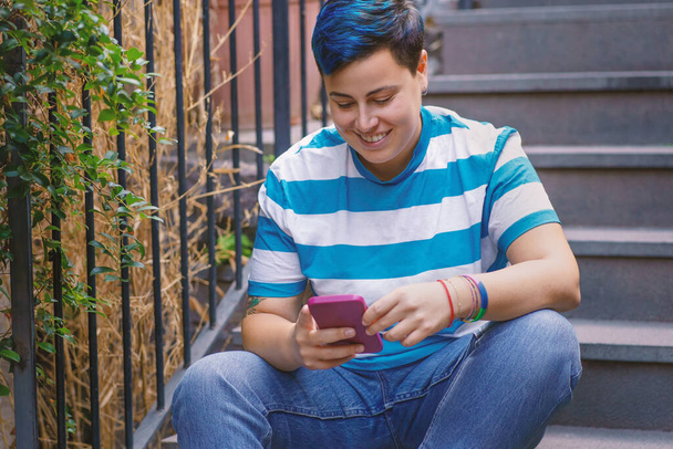 Persona no binaria vestida a rayas con smartphone, cómodamente sentada en escalones urbanos. - Foto, Imagen