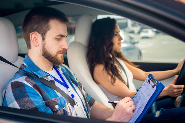 Инструктор мужского пола пишет заметки в тестовом отчете, в то время как молодая леди за рулем машины, смотрит на дорогу. Тест-драйв, транспорт, безопасность, концепция обучения - Фото, изображение
