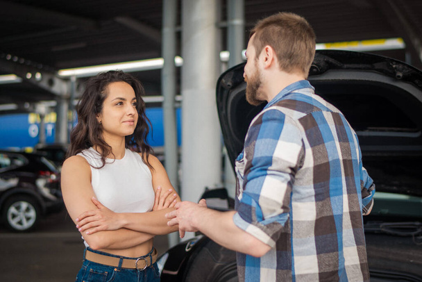 Young man explaining to a woman the reason for a car breakdown, hood open. Car service, insurance, transportation concept - Photo, Image