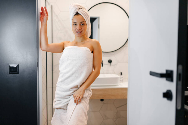 Jongedame met witte handdoek op het hoofd staande in de badkamer in de buurt van de grote wastafel en spiegel. Een vrouw gewikkeld in een handdoek. Levensstijl concept - Foto, afbeelding