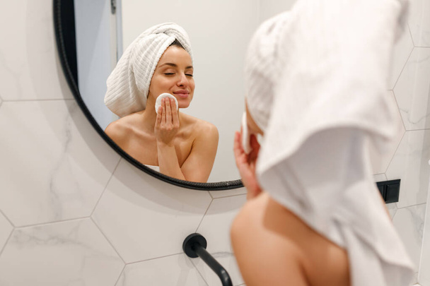 Mujer joven con toalla blanca en la cabeza de pie cerca del gran espejo en el baño. Concéntrate en el reflejo en el espejo. Concepto de estilo de vida - Foto, imagen