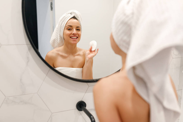 Glimlachende vrouw gehuld in witte handdoek kijkend in de grote spiegel terwijl ze in de badkamer stond. Routine, zelfzorg, lifestyle concept - Foto, afbeelding