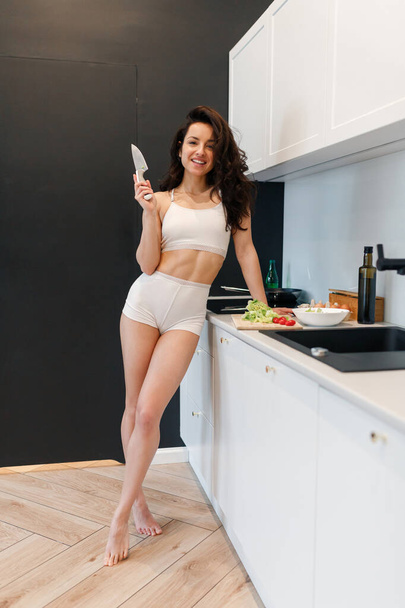 Full-length photo of young woman in white lingerie standing near the big kitchen table of the apartment. Lady holding a kitchen knife - Fotografie, Obrázek