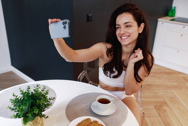 Lachende mooie dame in ondergoed die een telefoon vasthoudt terwijl ze foto 's maakt en' s morgens thuis thee drinkt. Lifestyle, ochtend routine concept. - Foto, afbeelding