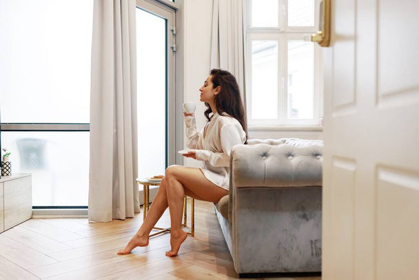 Ευτυχισμένη νεαρή γυναίκα με λευκό μεταξωτό πουκάμισο που αναπαύεται στον καναπέ στο διαμέρισμά της απολαμβάνοντας ζεστό ρόφημα το πρωί. Πλευρική άποψη. Τρόπος ζωής, έννοια ρουτίνα πρωί. - Φωτογραφία, εικόνα