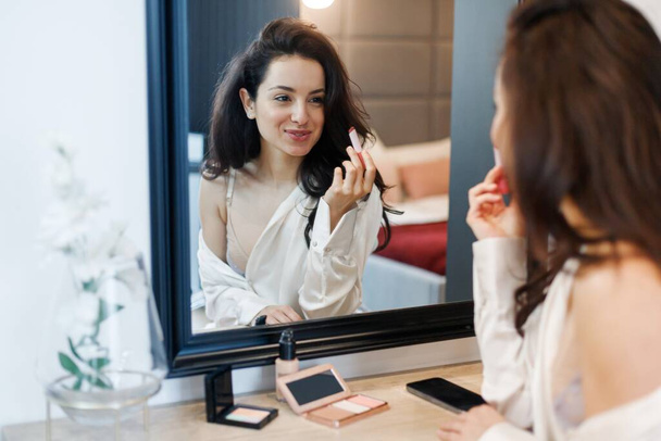 Ευτυχισμένη σέξι γυναίκα κάνει μακιγιάζ και τη χρήση κραγιόν, ενώ κάθεται μπροστά από τον καθρέφτη στο δωμάτιο. Ομορφιά και καλλυντικά προσώπου, έννοια ρουτίνα πρωί. - Φωτογραφία, εικόνα