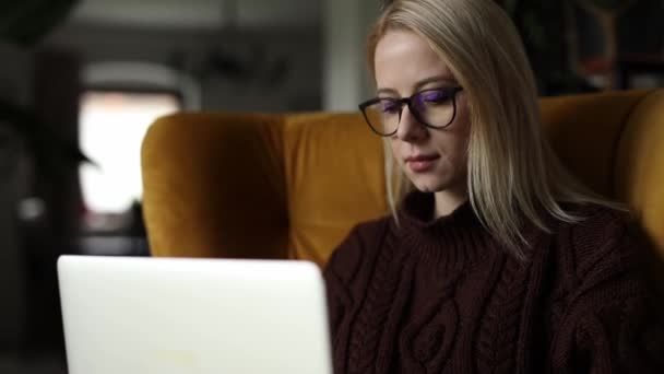Kaukasierin in Brille und Pullover mit Laptop sitzt zu Hause im Sessel - Filmmaterial, Video