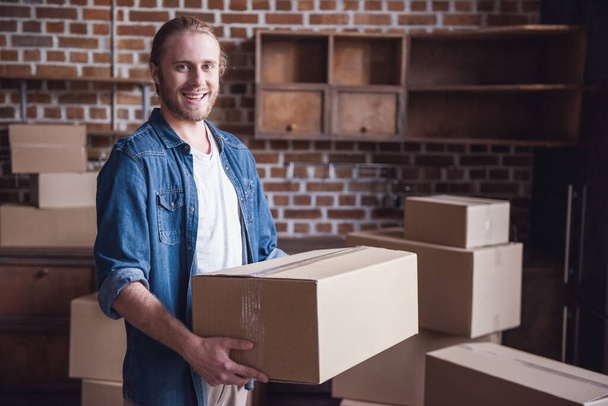 Handsome guy is holding a cardboard box, looking at camera and smiling while moving into new apartment - Foto, Imagen