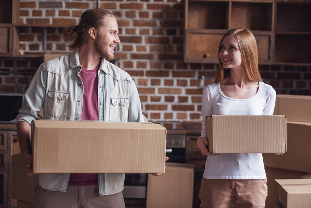 Красивая молодая пара держит картонные коробки, смотрит друг на друга и улыбается, переезжая в новую квартиру
 - Фото, изображение