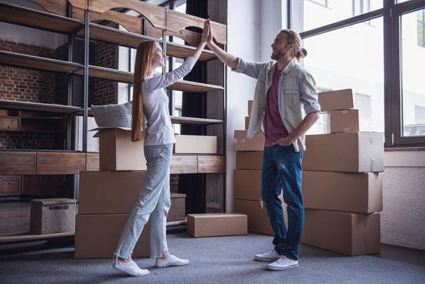 Full length portrait of beautiful young couple giving high five standing among cardboard boxes and smiling while moving into new apartment - Photo, Image