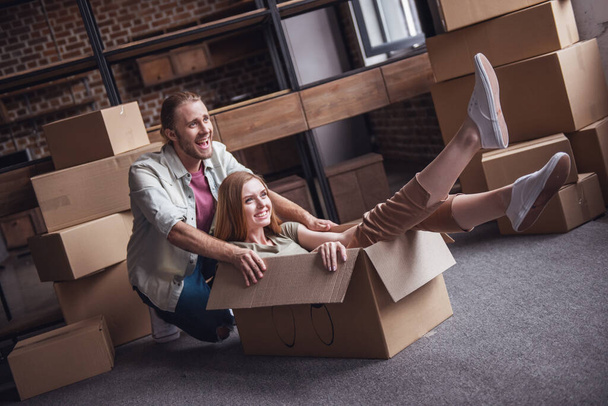 Beautiful young couple is having fun while moving into new apartment. Girl is sitting in the cardboard boxes - Photo, Image