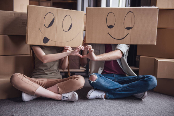 Gyönyörű fiatal pár ül a földön, az új lakás karton dobozok a fejükre, és mutatja a szív - Fotó, kép