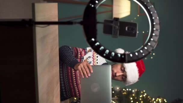 Arabische mannelijke blogger dragen Santa hoed unboxing pakket met nieuwe laptop computer op camera tijdens het opnemen van video-inhoud voor tech blog thuis op kerstavond, met behulp van smartphone op statief. Verticale video - Video