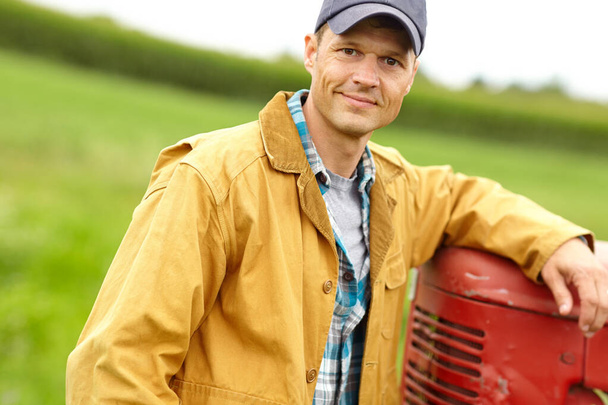 Tomando un descanso. Retrato de un granjero sonriente con el brazo apoyado en la capucha de su tractor mientras está de pie en un campo abierto - Foto, Imagen