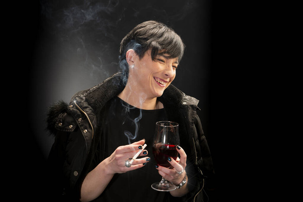 A young woman drinks wine and smokes a cigarette on a black background - Photo, Image