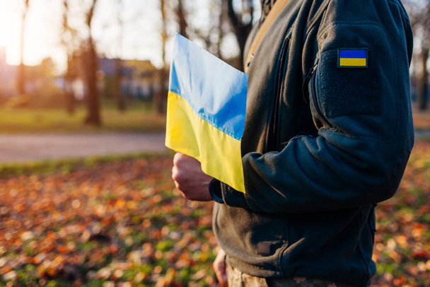 Κοντινό πλάνο της ουκρανικής σημαίας στο χέρι του στρατιώτη. Ο αμυντικός φοράει στρατιωτική στολή με ακίδα στην ύπαιθρο. Πόλεμος στην Ουκρανία. Ο άνθρωπος αγωνίζεται για ελευθερία ανεξαρτησία και ειρήνη - Φωτογραφία, εικόνα
