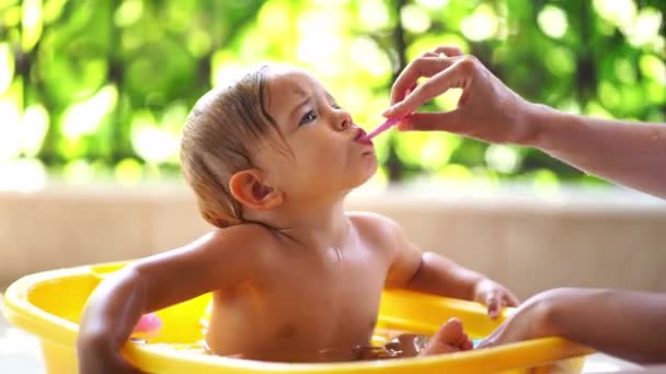 Mama putzt einem kleinen Mädchen, das in einer Schüssel mit Wasser sitzt, die Zähne. Hochwertiges 4k Filmmaterial - Filmmaterial, Video