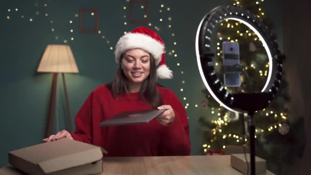 Női blogger rögzíti a videó tartalmat a modern okostelefonra, miközben kibontja laptop számítógép otthon karácsonykor. Befolyásoló felülvizsgálatot végez a modern kütyün. Fénymásolási hely - Felvétel, videó