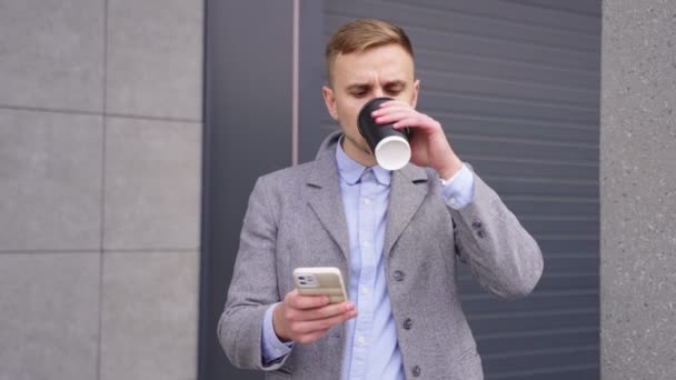 Νεαρός άνδρας πίνει καφέ και χρησιμοποιεί smartphone ενώ στέκεται κοντά σε κτίριο σε εξωτερικούς χώρους. Επιχείρηση, τεχνολογική έννοια. Αργή κίνηση - Πλάνα, βίντεο