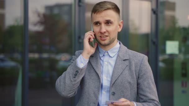 Hombre de negocios sonriente sosteniendo tarjeta de visita mientras habla teléfono inteligente, de pie al aire libre. Negocios, concepto tecnológico. Movimiento lento - Imágenes, Vídeo
