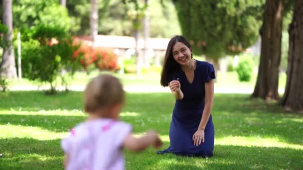 小さな女の子は、お母さんが彼女に花を持っている緑の芝生に沿って歩いていきます。高品質4k映像 - 映像、動画