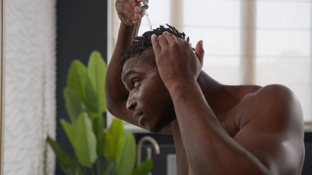 Afro-Amerikaanse man toepassen van arganolie serum op haarwortels dreadlocks gezonde massage ochtend badkamer routine man druppelen keratine cosmetische vloeistof behandeling kaalheid alopecia schoonheid zorg voor kapsel - Foto, afbeelding