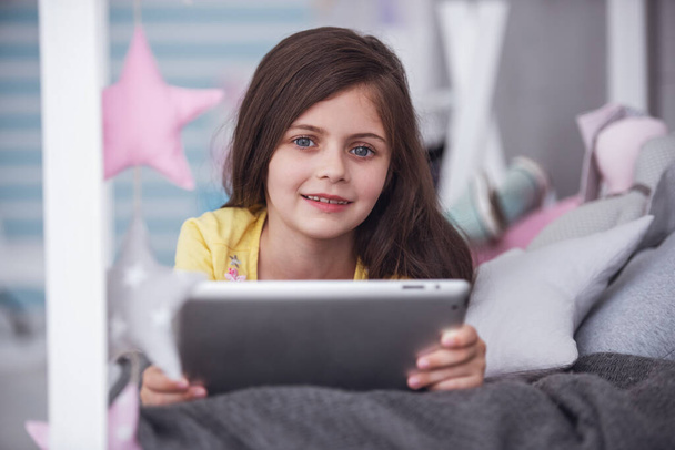Nettes kleines Mädchen benutzt ein digitales Tablet, blickt in die Kamera und lächelt, während sie zu Hause auf dem Bett liegt - Foto, Bild