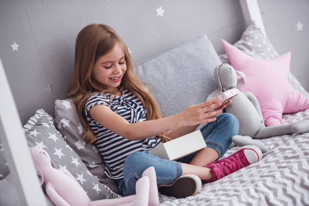 hübsches kleines Mädchen öffnet eine Geschenkbox und lächelt, während sie zu Hause auf ihrem Bett in ihrem Zimmer sitzt - Foto, Bild