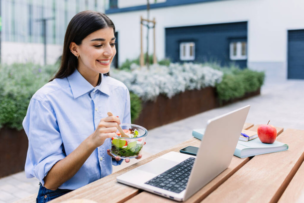Happy woman holding fresh salad while working on laptop sitting outside building office, looking at screen, watching video or listening colleague on video call. Business concept. - Photo, Image