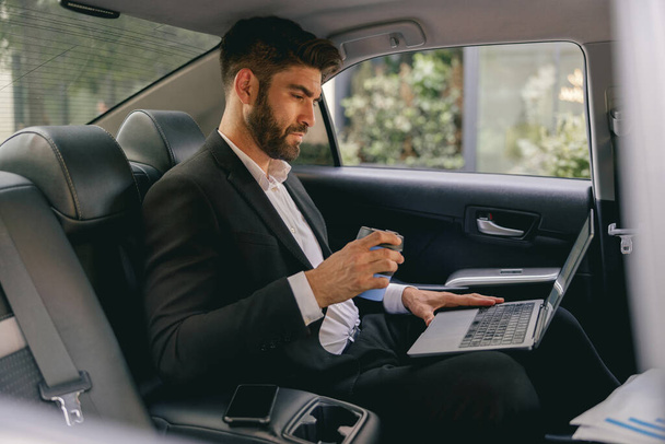 Professional male finansist working on laptop sitting car backseat and drinking coffee - Photo, Image