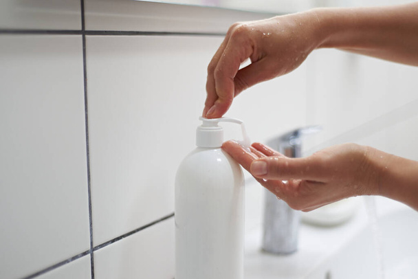 Рука крупным планом женщины сжимает жидкое мыло во время мытья рук, стоя у раковины в белой домашней ванной. Концепция очистки и гигиены. Здоровый образ жизни. Использование антисептического очищающего средства - Фото, изображение