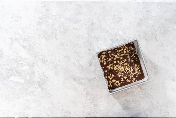 Flat lay. Filling square cheesecake pan lined with parchment paper with fudge mixture to prepare chocolate hazelnut fudge. - Photo, Image