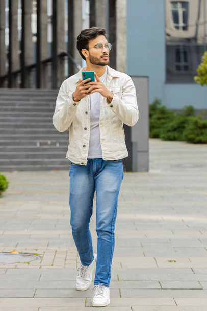 Jonge glimlachende Indiase man met behulp van smartphone typen sms-berichten, e-mail lezen, browsen nieuws buiten. Arabian Hindoe kerel met behulp van mobiele social media app lopen in het centrum van de stad straat. Zakenmensen - Foto, afbeelding