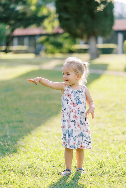 Das kleine lächelnde Mädchen steht auf einer sonnigen Wiese und zeigt mit der Hand nach vorne. Hochwertiges Foto - Foto, Bild