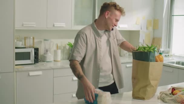 Nuori mies purkaa ostoksia paperipussista ja puhuu matkapuhelimella keittiössä päivällä kotona - Materiaali, video