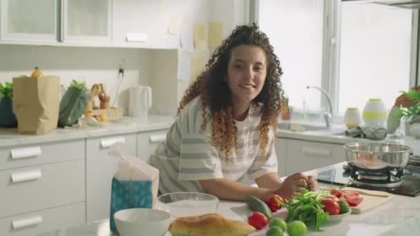 Μέτριο πλάνο χαρούμενα σγουρά μαλλιά κορίτσι κλίνει στο τραπέζι της κουζίνας με συστατικά τροφίμων και ποζάρουν για την κάμερα με χαμόγελο κατά τη διάρκεια της ημέρας στο σπίτι - Πλάνα, βίντεο