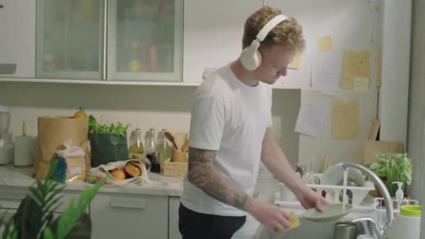 Νεαρός άνδρας ακούει μουσική με ασύρματα ακουστικά και χαμογελά ενώ πλένει πιάτα στην κουζίνα στο σπίτι - Πλάνα, βίντεο