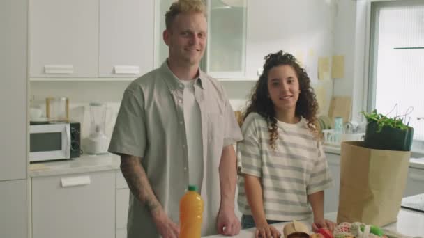 Média foto de jovem casal feliz em pé junto à mesa da cozinha com mantimentos, sorrindo e posando para câmera em casa - Filmagem, Vídeo