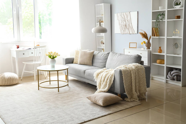Accogliente divano grigio con morbida coperta e tavolo all'interno del luminoso soggiorno - Foto, immagini
