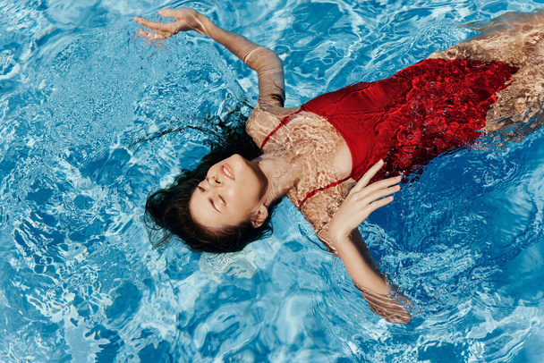 Mujer joven en la piscina en un traje de baño rojo con una hermosa sonrisa tumbada en el agua y nadando al sol nadando en la piscina, el concepto de relajarse en vacaciones. Foto de alta calidad - Foto, imagen