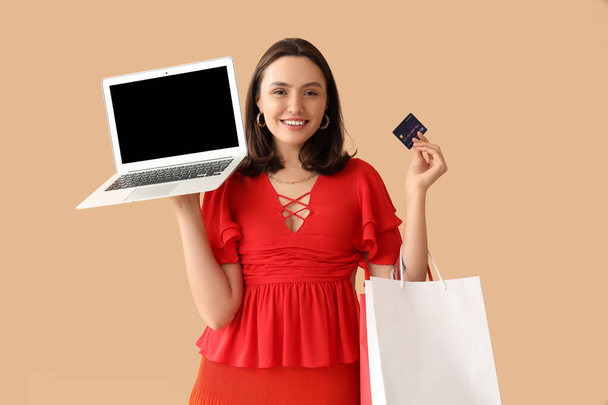 Kredi kartı, dizüstü bilgisayar ve bej arka planda alışveriş torbaları olan genç bir kadın. - Fotoğraf, Görsel
