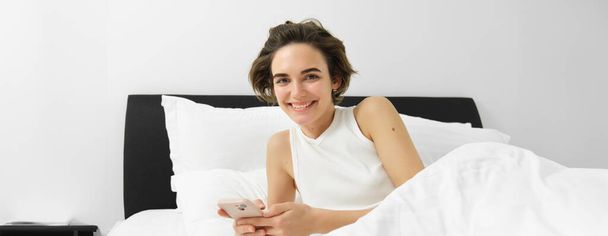 Immagine di una donna che controlla la sua app del sonno sul cellulare, sdraiata a letto e sorridente, usando lo smartphone in camera da letto. - Foto, immagini
