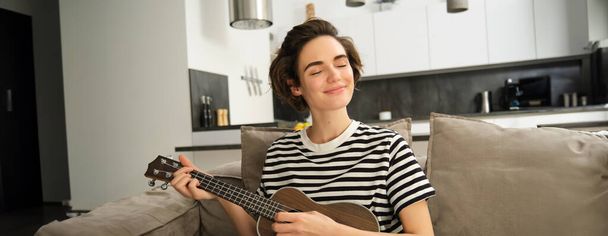 Detail portrét krásné usmívající se ženy, hraje ukulele, brnkání struny se zavřenýma očima a potěšenou tváří, sedí v obývacím pokoji. - Fotografie, Obrázek