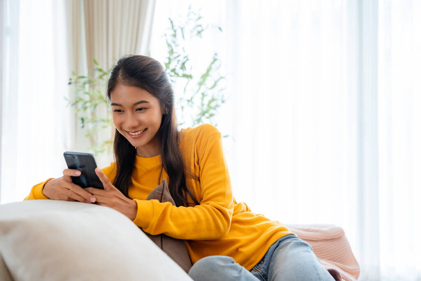 Happy young Asian woman sitting on sofa holding mobile phone using cellphone technology doing ecommerce shopping, buying online, texting messages relaxing on couch in cozy living room at home - Valokuva, kuva