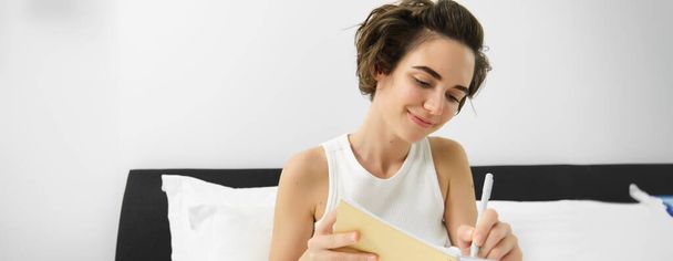 Portrait of young woman in bed, working or studying, writing in her journal, reading notebook notes, sharing thoughts with diary and smiling. - Photo, Image