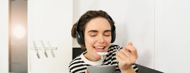 Κοντινό πορτρέτο της νεαρής γυναίκας απολαμβάνει να τρώει το πρωινό της, να τρώει δημητριακά με γάλα, να κρατάει κουτάλι και μπολ, να κάθεται στον πάγκο της κουζίνας με ακουστικά, να ακούει μουσική. - Φωτογραφία, εικόνα