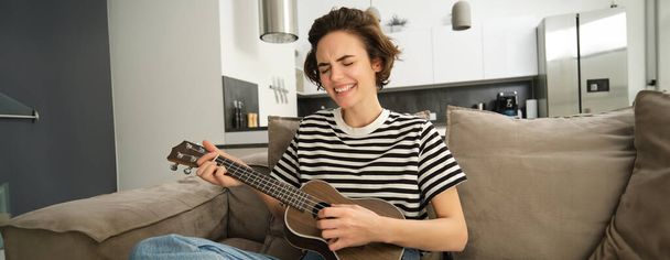 Retrato de una joven mujer moderna, estudiante tocando ukelele en casa, sentada con una pequeña guitarra, cantando y sintiéndose feliz, sentada en un sofá. Concepto de estilo de vida y música - Foto, imagen
