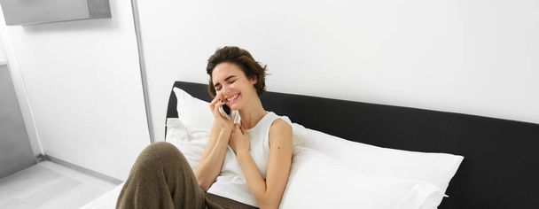 Weibliches Model sitzt mit Telefon im Bett, ruft Freundin an, telefoniert, lacht und lächelt. - Foto, Bild